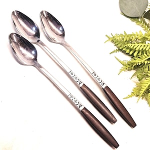 May include: Four silver spoons with dark brown wooden handles. The handles have a decorative silver and black geometric pattern. The spoons are arranged diagonally on a white surface, with greenery in the background.