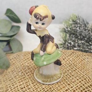 Może przedstawiać: Ceramiczna figurka siedzącego elfa z biedronką na głowie. Elf ma na sobie brązowo-żółty strój i siedzi na zielonym grzybie z białymi kropkami. Figurka ma kapryśną, vintage estetykę.