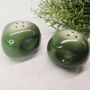Puede incluir: Un par de saleros y pimenteros de cerámica verde brillante y redondeada. Cada pieza presenta una forma lisa y bulbosa con una hendidura distintiva en el lateral y una superficie superior plana con cuatro pequeños orificios.