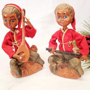 May include: Two vintage figurines of musicians. Each doll wears a red outfit with gold trim and a matching cap. One doll holds a stringed instrument, the other a flute. The dolls are seated on a brown and green base.