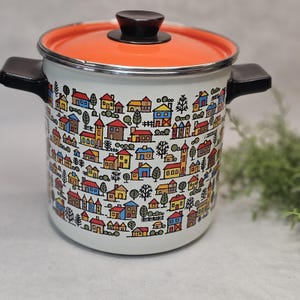 May include: A white enamel stockpot with a vibrant orange lid and black handles. The exterior features a repeating pattern of colourful houses, trees, and church steeples in shades of blue, yellow, and red against a white background.
