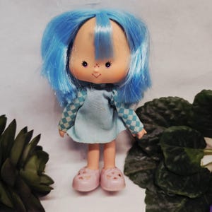 May include: A vintage doll with blue hair and a light blue dress. The doll has a smiling face with freckles and pink shoes. The dress features checkered sleeves. A collectible toy.