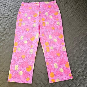 Puede incluir: Pantalones rosas con un estampado repetido de monos rosas, orbes amarillos y rectángulos crema. Los pantalones tienen un corte de pierna recta y cierre de botón y cremallera.
