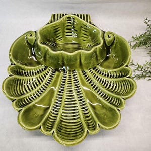 May include: A green ceramic serving dish in the shape of a seashell. The dish has a central bowl and a large, fan-shaped outer section with ribbed detailing. The glossy finish and olive green color give it a vintage aesthetic.