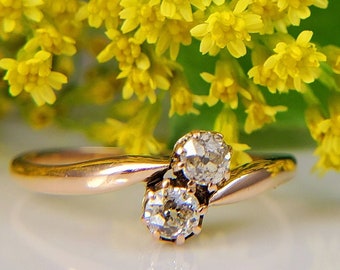 May include: A delicate gold ring with two round diamonds set in a twisted band. The ring is photographed against a backdrop of yellow flowers.