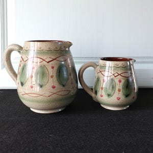 Peut inclure: Deux pichets en céramique avec des anses. Les pichets sont beiges avec des motifs de feuilles vertes et de points rouges. Les bords et l'intérieur sont bruns. Le plus grand pichet mesure environ 18 cm de haut, et le plus petit environ 10 cm.