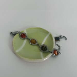 May include: A silver-toned bracelet with oval-shaped, multi-colored stones, resting on a small, light green ceramic dish. The stones are in shades of red, orange, yellow, and green. The dish has a white rim and a white line design.