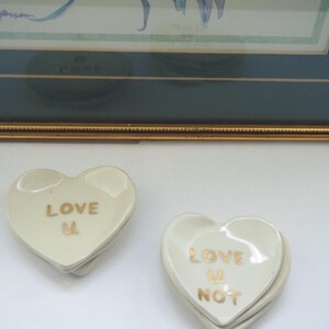 May include: Two stacks of heart-shaped ceramic dishes with a glossy off-white finish. The left stack features the gold text LOVE U, while the right stack displays the gold text LOVE U NOT. Each dish has a smooth, rounded edge and a concave center.