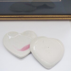 May include: Two heart-shaped, off-white ceramic dishes. One has a pink streak and a crack, the other has faint markings. The dishes are on a white surface, with a framed picture in the background.
