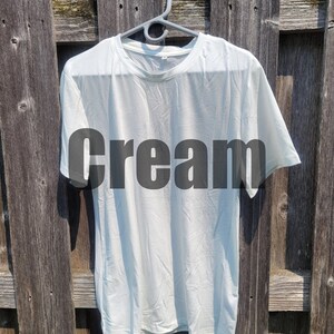May include: A plain white t-shirt hanging on a hanger against a wooden background. The word "cream" is printed in black on the front of the shirt.