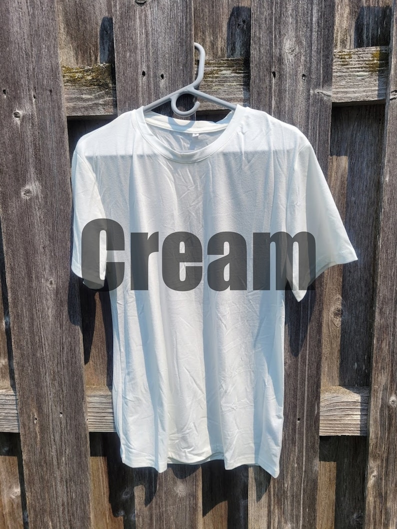 May include: A plain white t-shirt hanging on a hanger against a wooden background. The word "cream" is printed in black on the front of the shirt.