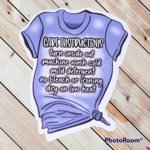 Peut inclure: Un t-shirt violet avec des instructions d'entretien imprim&eacute;es dessus. Les instructions disent : "Retourner &agrave; l'envers, laver en machine &agrave; froid, d&eacute;tergent doux, pas de javel ni de repassage, s&eacute;cher &agrave; basse temp&eacute;rature."