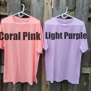 May include: Two plain t-shirts hanging on hangers against a wooden fence. The t-shirt on the left is coral pink and says "Coral Pink". The t-shirt on the right is light purple and says "Light Purple".