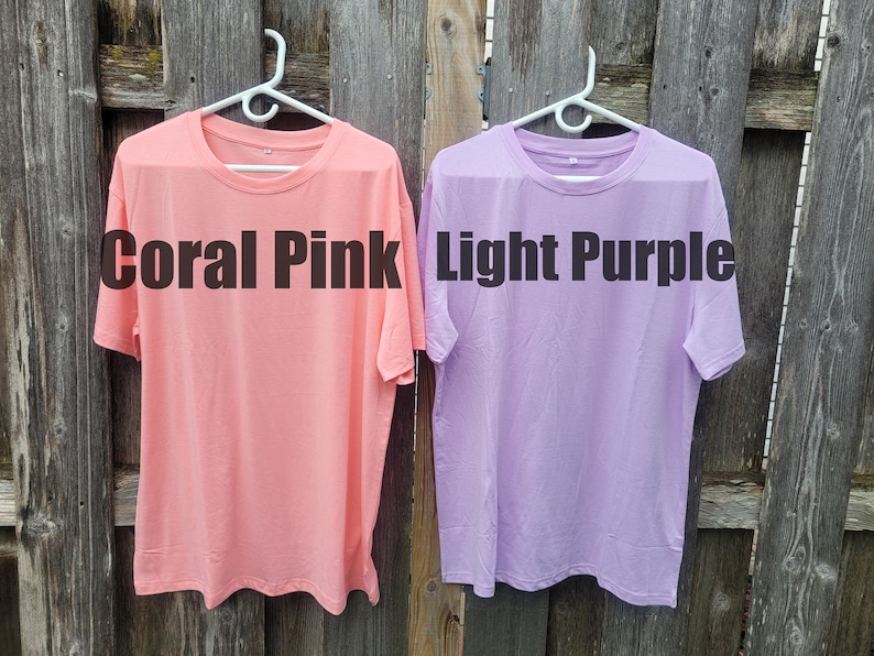 May include: Two plain t-shirts hanging on hangers against a wooden fence. The t-shirt on the left is coral pink and says "Coral Pink". The t-shirt on the right is light purple and says "Light Purple".