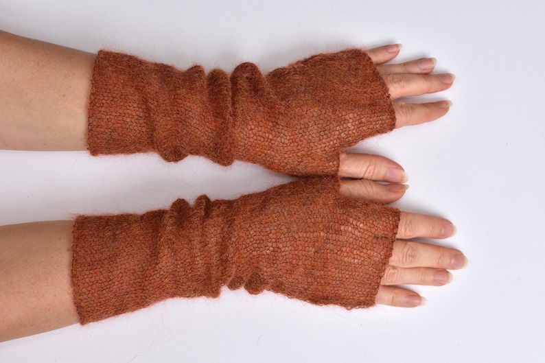 May include: A pair of rust-colored fingerless gloves made from a lightweight, semi-sheer knitted fabric. The gloves feature a textured, loose-knit construction that extends from the knuckles to the mid-forearm, with a simple opening for the thumb.