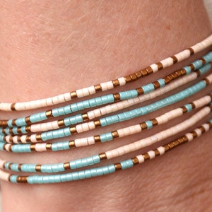 May include: A stack of beaded bracelets in shades of white, light blue, and gold. The bracelets are made of small, round beads and are layered on top of each other.