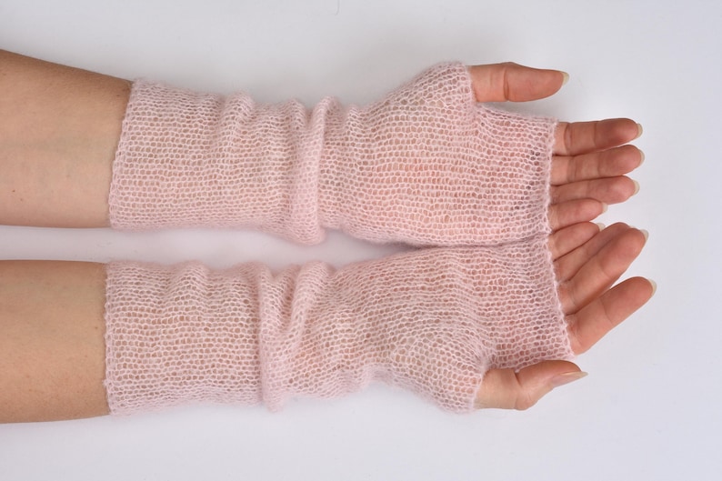 May include: A pair of pale pink fingerless gloves crafted from a fine, sheer, knitted yarn. These arm warmers feature a delicate, open-stitch texture that extends from the knuckles up to the mid-forearm, providing a lightweight layer.