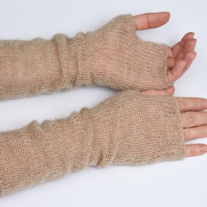 May include: A pair of beige fingerless gloves made from a sheer, loosely knitted yarn. The gloves extend from the knuckles to the mid-forearm, featuring a soft, textured appearance with a visible open-weave pattern throughout the fabric.
