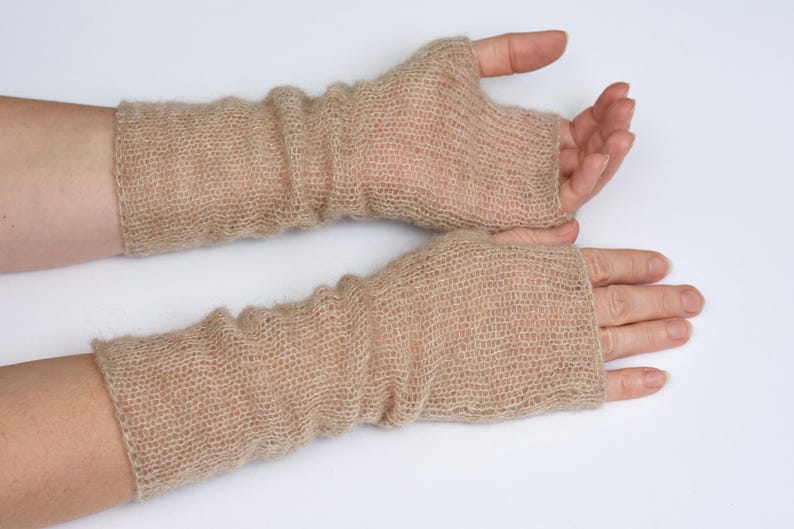 May include: A pair of beige fingerless gloves made from a sheer, loosely knitted yarn. The gloves extend from the knuckles to the mid-forearm, featuring a soft, textured appearance with a visible open-weave pattern throughout the fabric.