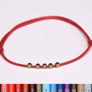 Puede incluir: Un brazalete de cuerda roja con cuentas cuadradas doradas. El brazalete está atado con un nudo en el extremo.