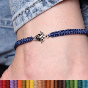 Può includere: Un braccialetto intrecciato blu navy con un ciondolo a forma di tartaruga argentata. Il braccialetto è mostrato sul polso di una persona.