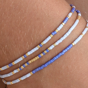 Pode incluir: Três pulseiras sobrepostas com contas brancas, azul claro e azul escuro intercaladas com contas douradas.