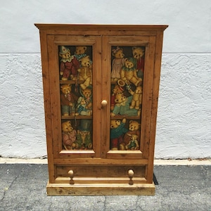 May include: Wooden cabinet with two glass doors, featuring a collection of teddy bears in various outfits. The cabinet has a drawer with two wooden knobs and a light brown finish. A charming piece for a child's room or a collector.