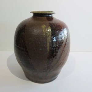 Puede incluir: Un jarrón de cerámica grande, de color marrón oscuro, con un cuerpo redondeado y un cuello estrecho. El jarrón tiene una superficie texturizada con variaciones de color, incluyendo marrón oscuro, marrón rojizo y toques dorados. El borde es marrón claro.
