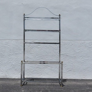 May include: A tall, chrome-finished shelving unit with four glass shelves. The unit has a curved top and gold-colored accents on the corners. The shelves are rectangular and the frame is made of metal.