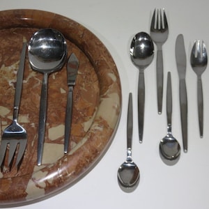 May include: A collection of stainless steel silverware, including forks, spoons, and knives, arranged on a decorative marble tray. The silverware has a simple, modern design with long, slender handles. The tray has a brown and beige speckled pattern.