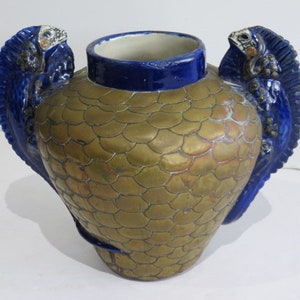 Könnte beinhalten: Eine dekorative Keramikvase mit einem goldfarbenen, schuppenartigen Muster auf dem Körper. Die Vase hat einen blauen Rand und zwei blaue, flügelartige Griffe mit reptilienartigen Figuren. Der Innenraum ist weiß.