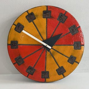 May include: A round, decorative wall clock with a segmented design in orange and yellow. The clock face features dark brown square markers and black hands. The clock's diameter is approximately 12 inches.
