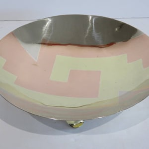Peut inclure: Un bol décoratif en métal avec un motif géométrique. Le bol a un bord argenté et un centre avec un motif de formes roses et jaunes. Le bol a un petit pied doré.