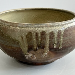 May include: A round ceramic bowl with a wide opening and a curved body. The exterior features a dark brown base colour with a thick, textured cream-coloured glaze dripping from the rim down the sides, creating a layered and mottled appearance.
