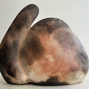 Op de afbeelding: Een keramische sculptuur met een afgeronde basis en een omhoog gebogen, langwerpige vorm. Het stuk heeft een mix van kleuren, waaronder tinten bruin, zwart en roze, waardoor een textuurrijk, organisch uiterlijk ontstaat. De sculptuur is waarschijnlijk een decoratief kunstwerk.