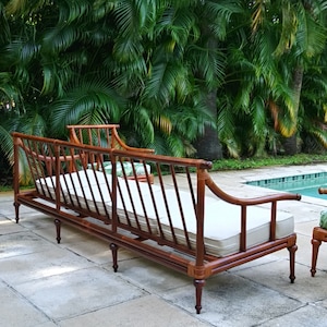 Könnte beinhalten: Ein hölzernes Outdoor-Sofa mit einem hellbeigen Kissen und einem passenden Stuhl. Das Sofa hat einen dunkelbraunen Rahmen mit einem gewebten Design und geschwungenen Armlehnen. Der Hintergrund zeigt üppiges grünes Laub und einen Swimmingpool.