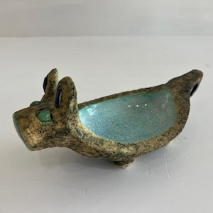 Puede incluir: Un plato de cerámica hecho a mano con forma de animal, posiblemente un conejo o un perro. El plato tiene un exterior marrón terroso texturizado con un interior turquesa. El animal tiene ojos verdes y detalles azules.