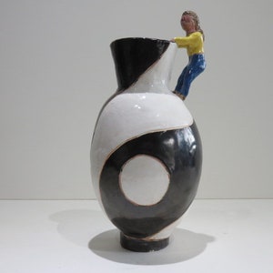 Może przedstawiać: Ceramiczny wazon z czarno-białym wzorem wirów i okrągłym środkiem. Do krawędzi przymocowana jest mała, rzeźbiona figurka w żółtej koszuli i niebieskich spodniach, która wygląda, jakby wspinała się na krawędź naczynia.