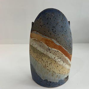 May include: A tall, rounded ceramic vase featuring a speckled blue glaze at the top and bottom, with diagonal bands of tan, cream, and rust-colored glaze across the center. The surface has a textured appearance with visible dark speckles throughout.