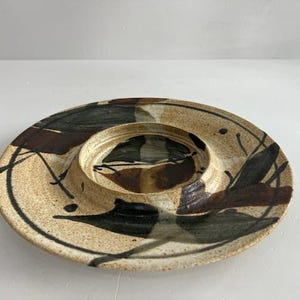 May include: A round ceramic serving dish with a recessed center, featuring a speckled beige base with abstract brushstrokes in dark brown and black. The surface has a glossy finish with visible glaze variations and organic, hand-painted markings.