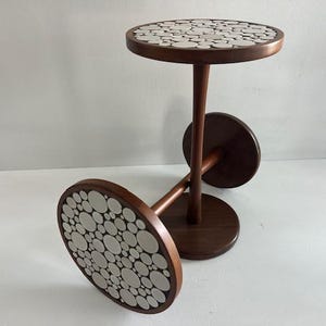 May include: A two-tiered side table with a dark wood frame. The round tabletop and base feature a mosaic design of white and grey circular tiles. The table has a mid-century modern aesthetic.