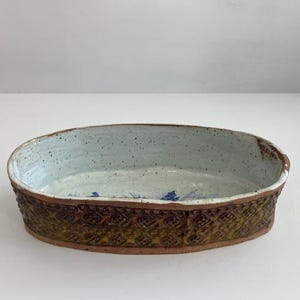 Puede incluir: Fuente ovalada de cerámica con exterior texturizado y estampado en tonos marrón y amarillo. El interior es azul claro con detalles moteados y detalles florales azules. Una pieza de cerámica hecha a mano.