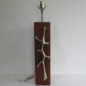 May include: A mid-century modern table lamp with a rectangular wooden base and a brushed metal accent. The lamp features a silver-colored, organic-shaped design on the front. The lamp has a silver-colored stem and a light bulb socket.