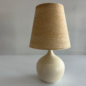May include: A table lamp featuring a smooth, off-white, spherical ceramic base and a tapered, cylindrical lampshade with a textured, light tan, horizontal fiber-like pattern. The lamp stands upright with a clean, simple silhouette and neutral tones.