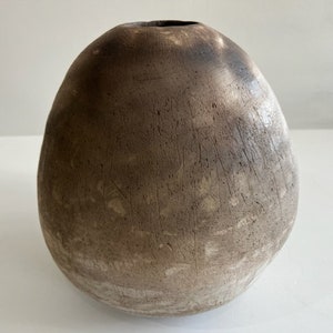 May include: A round, egg-shaped ceramic vase with a textured, matte surface. The exterior features a gradient of dark brown and charcoal tones at the top, transitioning into a lighter, dusty beige colour toward the base, with visible vertical markings.