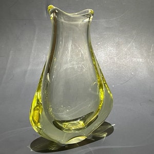 May include: A clear glass vase with a teardrop-shaped body and a yellow-tinted base. The vase features a narrow, undulating rim and thick, smooth glass walls that taper toward the top, creating a fluid, organic silhouette with a hollow center.