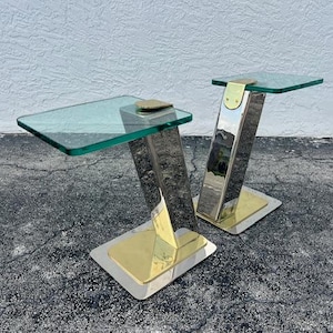 Puede incluir: Dos mesas auxiliares modernas con tapas de vidrio transparente y bases metálicas. Las bases presentan una combinación de metal plateado y dorado, creando un contraste llamativo. Las mesas tienen un diseño angular único, ideal para un espacio de vida contemporáneo.