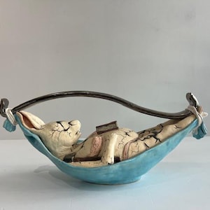 Puede incluir: Una escultura de cerámica de un cerdo relajándose en una hamaca azul. El cerdo es de color crema con grietas oscuras, y un pequeño libro descansa sobre su vientre. La hamaca está sostenida por una barra de metal curvada y atada con detalles azules.