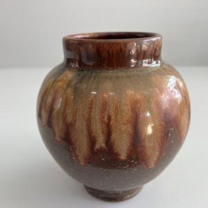Puede incluir: Un pequeño jarrón redondo de cerámica con borde y base marrón oscuro. El cuerpo del jarrón tiene una mezcla de tonos marrones, tostados y verdes, con un efecto de esmalte goteado. El jarrón es un objeto decorativo.