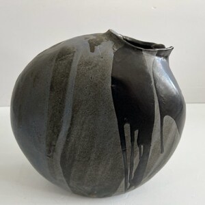 Può includere: Un vaso in ceramica rotondo e asimmetrico con una finitura maculata grigio scuro e nera. La superficie presenta uno smalto nero lucido che cola lungo il lato, in contrasto con il corpo grigio opaco e testurizzato e una piccola apertura.